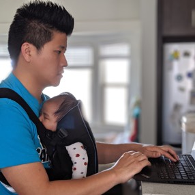 Father typing with a child in a front-facing baby carrier