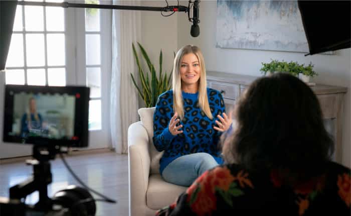Justine Ezarik, better known as iJustine, wearing jeans and a blue sweater with leopard print, speaks candidly with an interviewer who faces away from the camera. A microphone can be seen at the top of the frame and a video camera recording the interview takes up the left foreground.